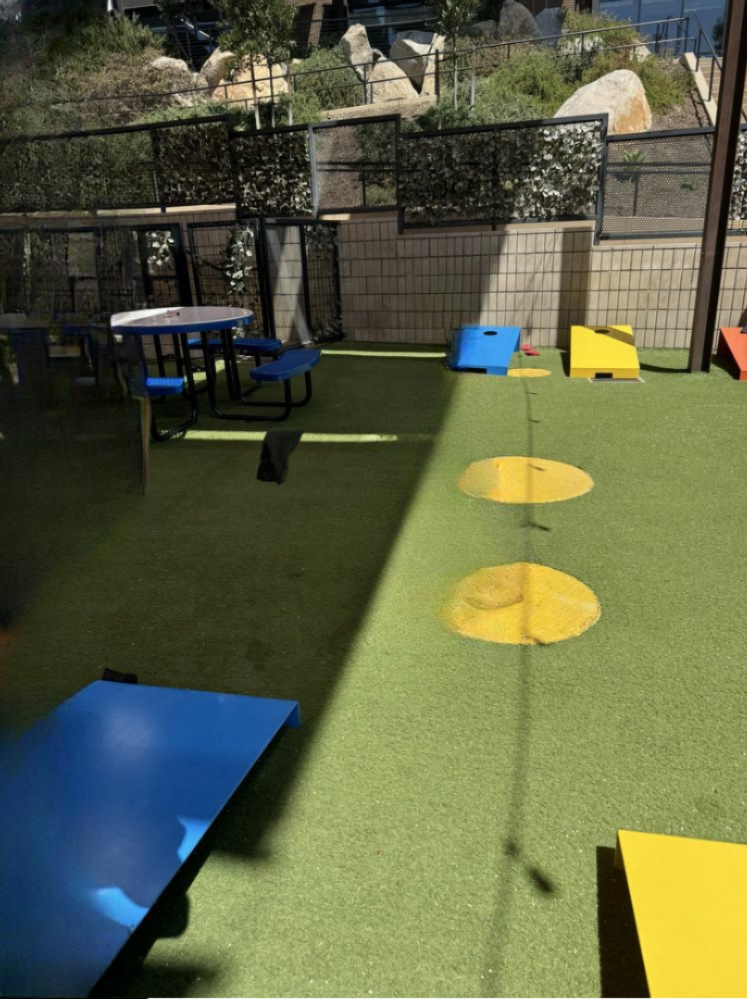 A bright blue and yellow corn hole setup on artificial turf, surrounded by picnic tables under an open-air patio.