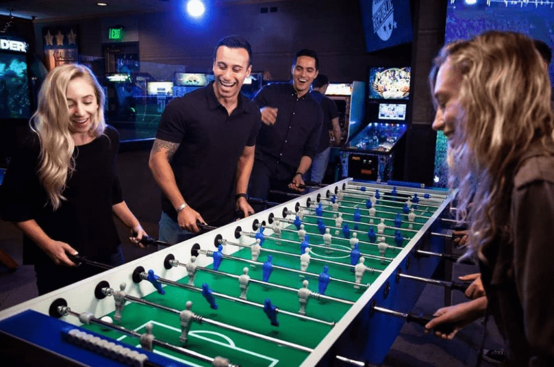 Friends enjoying a foosball game at Draft Republic San Marcos with arcade games in the background.
