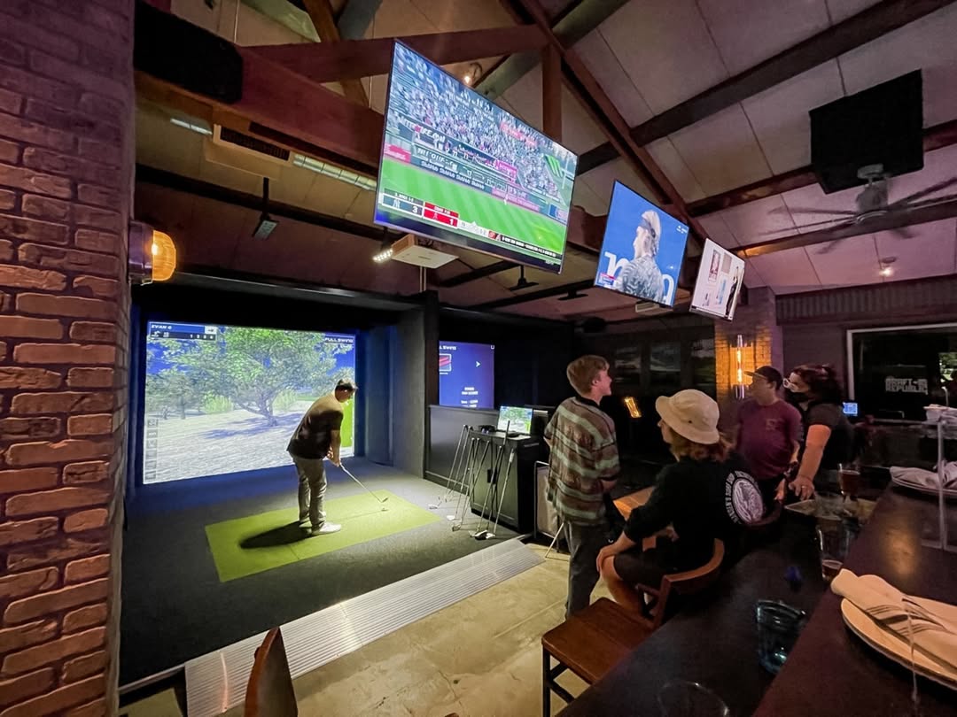 A group watches a player use the simulator, surrounded by multiple TV screens and bar seating.