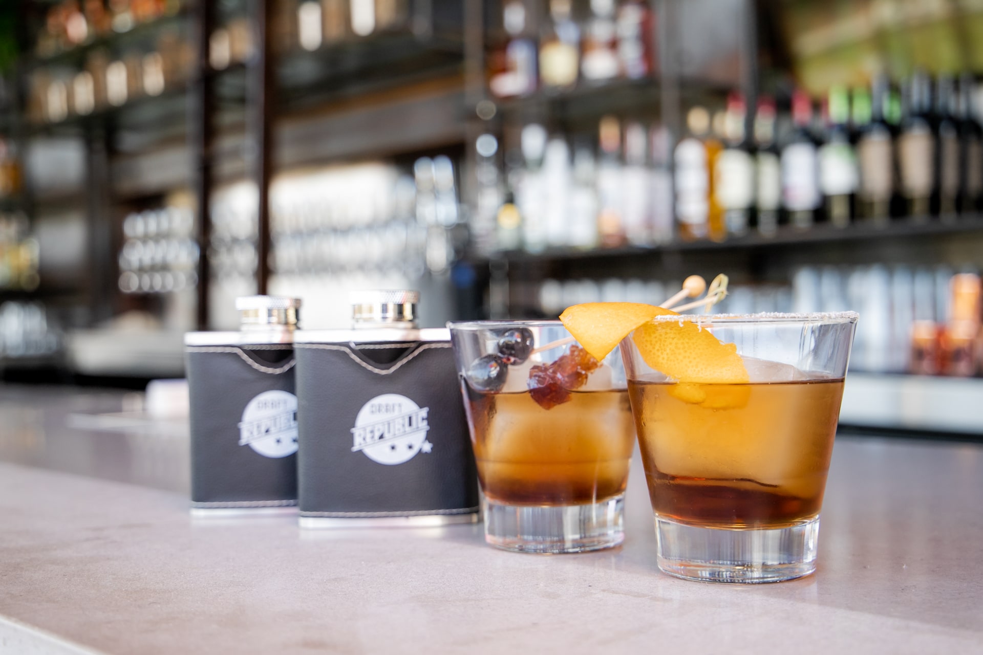 A stylish bar setting featuring handcrafted cocktails with garnishes, alongside branded flasks at a San Marcos happy hour spot.