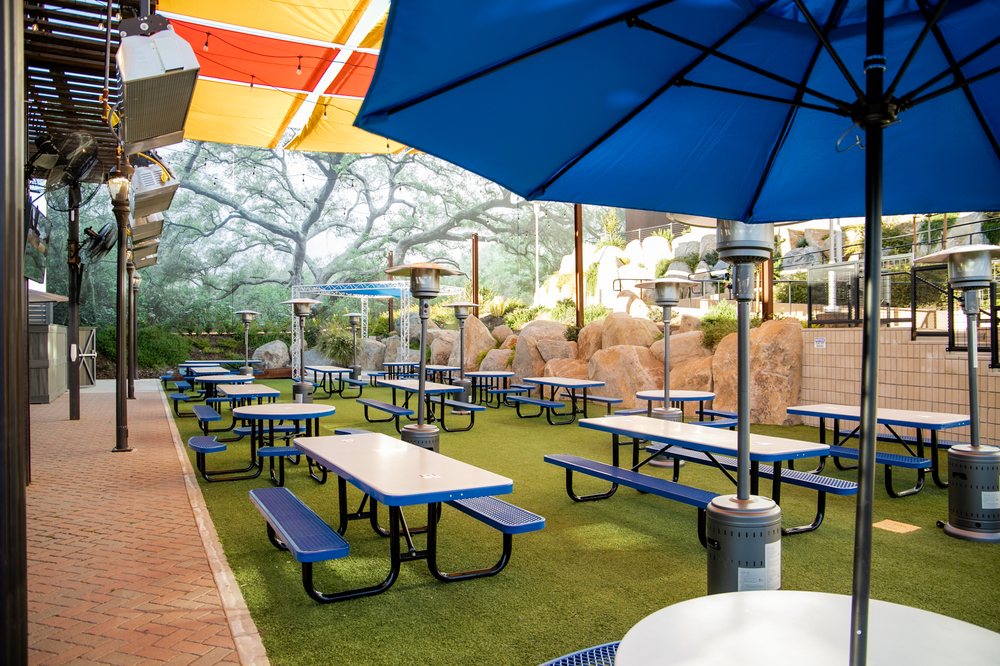 A lively patio area with shaded picnic tables, outdoor heaters, and ample space for games like corn hole.