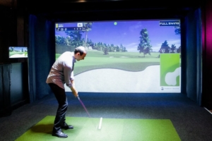 A player taking a shot at the simulator, with a high-definition screen displaying a virtual golf course.